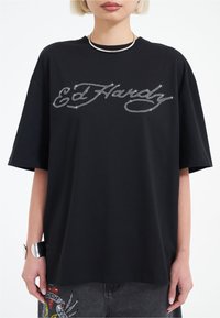 Camiseta de algodón negra con diseño de logo de strass. Manga corta, corte holgado y estilo cuello redondo. Textura suave.