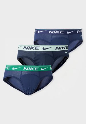 Modèle masculin debout pieds nus portant un slip Nike bleu foncé avec une ceinture bleu clair sur un fond gris clair uni.