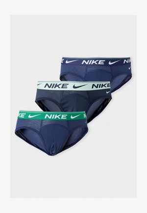 Modèle masculin debout pieds nus portant un slip Nike bleu foncé avec une ceinture bleu clair sur un fond gris clair uni.