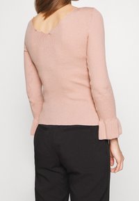 Hellrosa, geripptes Sweatshirt mit gewelltem Ausschnitt und ausgestellten Ärmeln, von hinten gezeigt, kombiniert mit schwarzen, taillierten Hosen.