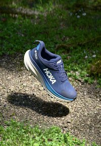 Blauwe HOKA hardloopschoen met een bovenwerk van mesh, een gevoerde witte zool en zwarte accenten. Voorzien van veters en textuurpatronen op het bovenwerk.