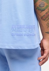 Light blue cotton t-shirt featuring embroidered text on the hem: "ESTABLISHED Milano 1971" and "COPYRIGHT @ Cola Cola." Soft texture, relaxed fit.