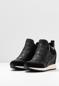 Svarta kilklack sneakers med kviltat läder, metalldekaler, siddragkedjor och vita sulor på en vit yta med grå bakgrund.