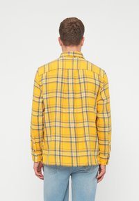 Gelbes karierter Hemd mit blauen und schwarzen Karomuster. Verfügt über Knopfmanschetten und ein yoke-Design. Getragen mit hellblauen Jeans.