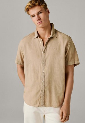 Joven vestido con una camisa beige de manga corta y pantalones blancos, de pie con las manos detrás de la espalda contra un fondo liso.