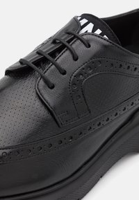 Scarpa in pelle nera con dettagli perforati, lacci piatti e suola testurizzata. Presenta una punta arrotondata e un rivestimento interno a contrasto.