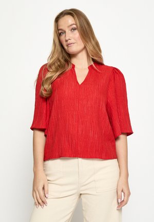 Rote, kurz geschnittene Bluse mit strukturierter Oberfläche, V-Ausschnitt und kurzen Puffärmeln, kombiniert mit hellbeigen Hosen.