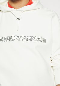 Vit huvtröja med en framträdande broderad "Emporio Armani"-logotyp i svart. Bomullstyg med en mjuk textur. Röd foder synlig.