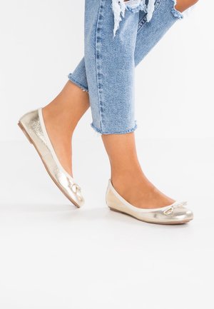 Ballerines dorées avec une finition brillante, des bordures blanches et un petit nœud. Portées avec un jean en denim déchiré cropped. Semelle plate.