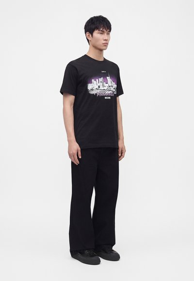 Jeune homme debout portant un t-shirt noir avec un graphisme de skyline de ville et un pantalon large noir sur fond blanc.