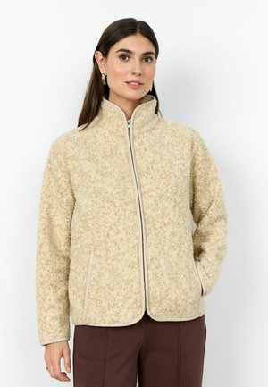 Kobieta ubrana w beżową, teksturowaną kurtkę na zamek z wysokim kołnierzem i bocznymi kieszeniami, w połączeniu z ciemnobrązowymi spodniami, stojąca na białym tle.