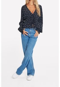 Marinebluse med hvide prikker, v-udskæring, indsnøret talje og lange ærmer, parret med højhalset blå jeans og hvide sneakers.