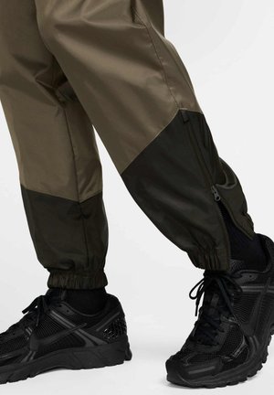 Pantalones de nailon verde oliva con detalles negros, que cuentan con puños ajustados, aberturas laterales con cremallera y se combinan con zapatillas deportivas negras.