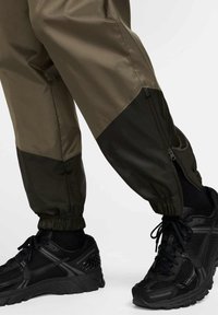 Olivově zelené a černé sportovní kalhoty s elastickými lemy a bočním zipem, doplněné černými sportovními botami s meshovým designem a strukturovanými detaily.