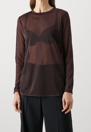 Person iført gennemsigtig brun langærmet top over sort BH og sorte bukser, stående mod en ensfarvet lys baggrund.