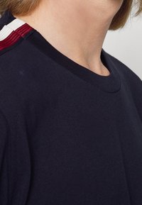 Granatowa koszulka z okrągłym dekoltem, z czerwoną i białą paskowaną aplikacją na ramieniu. Wykonana z miękkiego, gładkiego materiału.