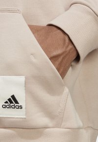 Vaaleanbeige collegepaita, jossa on ribbaaset hihansuut ja etutasku. Musta Adidas-logo-tasku etutaskussa. Sileä kangas.