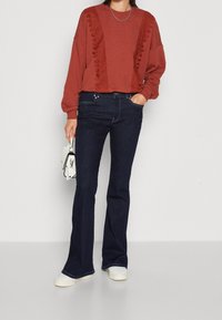 Červenohnědý krátký svetr s texturovanými detaily, tmavé džínové rozšířené džíny a bílé nazouvací boty, doplněné malou bílou kabelkou.