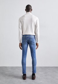 Světle šedý rolák, slim fit modré džíny, černé boty; pohled zezadu, ukazující přiléhavou siluetu a hladkou texturu látky.