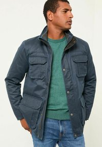 Marineblau, wasserdichte Jacke mit hohem Kragen, mehreren vorderen Taschen und Details aus Messing, über einem grünen Pullover getragen.