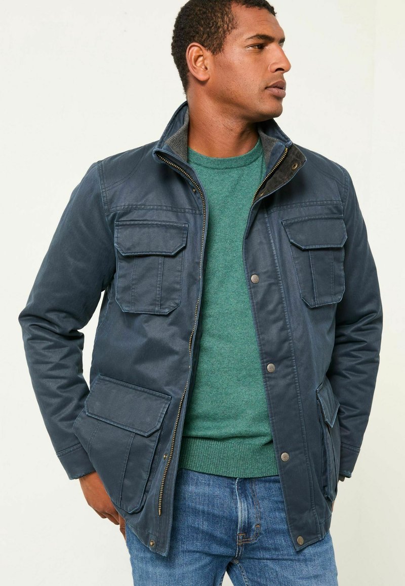 Marineblau, wasserdichte Jacke mit hohem Kragen, mehreren vorderen Taschen und Details aus Messing, über einem grünen Pullover getragen.