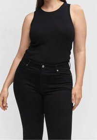 Nainen, joka on pukeutunut tiukkaan mustaan hihattomaan ribbipaitaan ja korkeavyötäröisiin mustiin housuihin, seisoo yksinkertaista vaaleaa taustaa vasten.