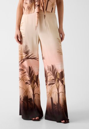 Kobieta w spodniach o szerokich nogawkach z gradientem od beżu do ciemnego brązu i sylwetkami palm, w połączeniu z pasującym topem wiązanym z przodu.