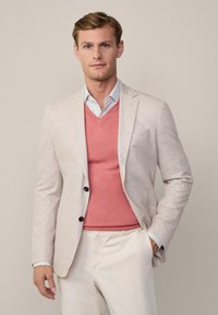 Sacou bej deschis cu două nasturi negri, purtat peste un pulover corai cu decolteu în V. Pantaloni albi slim-fit și o cămașă în dungi albastre deschise.