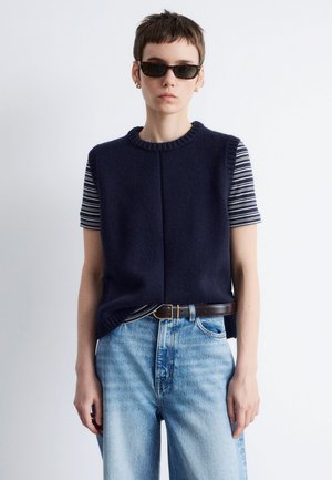 Granatowy dzianinowy sweter bez rękawów nałożony na koszulę w paski z krótkim rękawem, w połączeniu z jasnoniebieskimi dżinsami i brązowym paskiem. Całość dopełniają okulary przeciwsłoneczne.