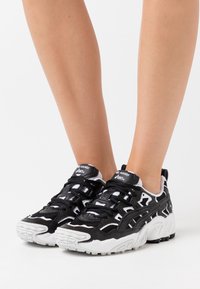 Svart och vit Asics Gel-Nandi sneakers som bärs på fötter, med kraftiga sulor och intrikata paneler mot en vit bakgrund.