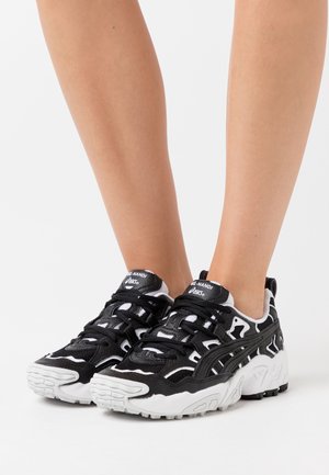 Černobílé tenisky Asics Gel-Nandi na nohou, vybavené robustními podrážkami a složitým designem panelů na bílém pozadí.