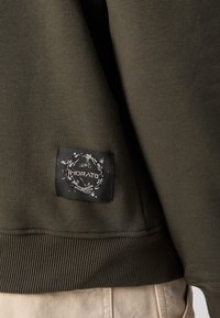 Sweatshirt vert foncé avec un ourlet côtelé et une étiquette brodée avec un motif floral, placée sur le bas à gauche.