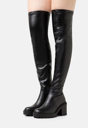 Botas negras por encima de la rodilla hechas de faux cuero brillante. Cuentan con un tacón grueso y una cremallera expuesta en el lado interno.