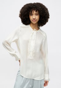 Weiße Bluse mit langen Ärmeln, die einen ausgefransten Ausschnitt und Saum hat, sowie einer Bindegarnitur an der Vorderseite. Hergestellt aus leichtem Stoff.