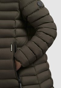 Padded olive green jacket featuring a zippered pocket and a round black logo patch on the shoulder. Textured, quilted design.