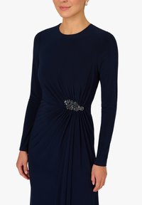 Vrouw draagt een marineblauwe jurk met lange mouwen, een geplooide taille en een donker bloemenbroche aan de rechterkant.