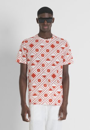 Camiseta blanca de manga corta con un patrón geométrico rojo, que presenta diseños triangulares y florales. La tela parece suave y ligera.