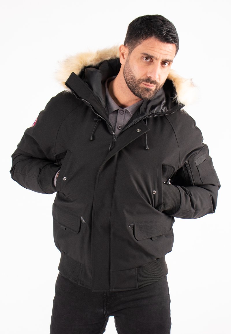 Veste noire à capuche avec bordure en fausse fourrure, équipée d'une fermeture éclair et de boutons-pression, poches avant et poignets côtelés. Le matériau semble lisse et résistant.