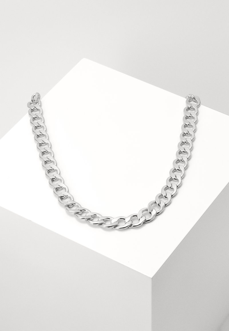 Collier chaîne épaisse en argent présenté sur un socle blanc incliné sur un fond gris clair.