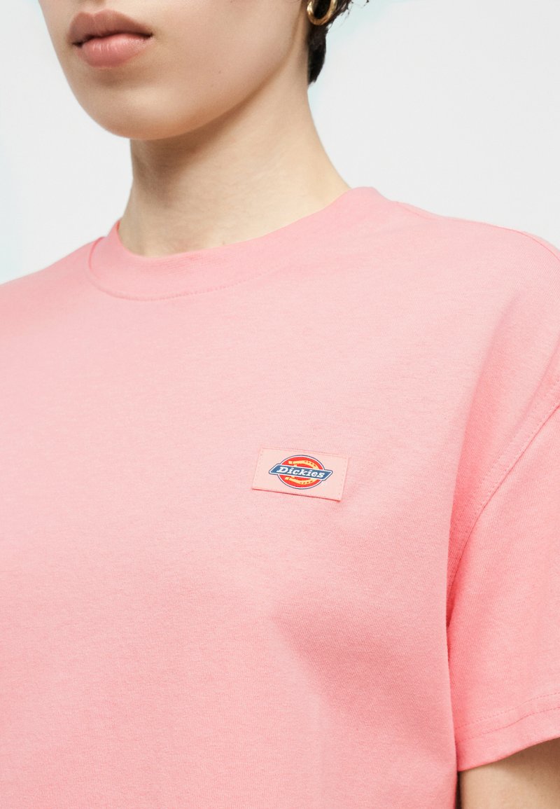 Close-up van een persoon die een roze T-shirt draagt met een kleine Dickies logo patch op de borst en gouden oorringen.
