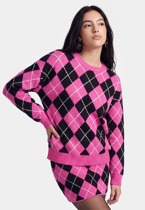 Femme aux longs cheveux foncés portant un pull motif losanges rose et noir et une jupe assortie, posant avec la main sur la hanche.