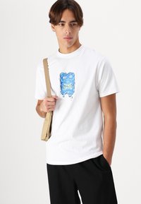 Hvid bomulds t-shirt med et farverigt grafisk motiv af et blåt ansigt med skarpe tænder. Modellen holder en lysebrun skuldertaske og har sorte bukser på.