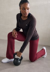 Melnā kettlebell, ko tur kneeling pozīcijā. Individuāls valkā brūnu garām piedurknēm topu un bordo izplatītus bikses, ar baltām kedām.