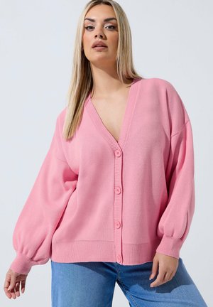 Vrouw draagt een loszittende roze cardigan met knopen en ballonmouwen, gecombineerd met een blauwe spijkerbroek, tegen een effen lichte achtergrond.