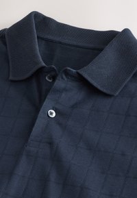 Temno-modra polo majica z reliefnim vzorcem v obliki diamanta, ki ima tradicionalni ovratnik, odprtino s tremi gumbi in gladek zaključek blaga.