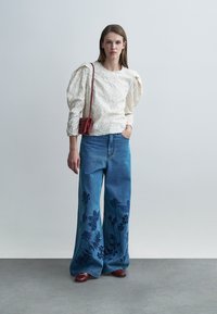 Κρεμ μπλούζα με υφή και φουσκωτά μανίκια, συνδυασμένη με μπλε τζιν παντελόνι φαρδιές μπατζάκια με σκούρες floral κεντητικές λεπτομέρειες. Μικρή κόκκινη τσάντα, καφέ παπούτσια.