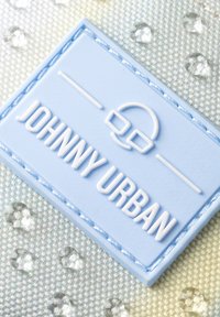 Etichetta in gomma blu chiaro con testo bianco in rilievo "JOHNNY URBAN" e grafica. Bordo cucito in blu su uno sfondo argentato a motivi.