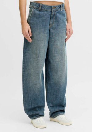 Person iført løstsiddende blå denimjeans og hvide sneakers, stående mod en ensfarvet lys baggrund.
