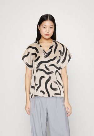 Kortærmet beige bluse med sort abstrakt penselstrøgsmønster, med V-udskæring og en løs, luftig pasform. Stylet med grå bukser.