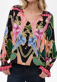 Kobieta w zapiętej bluzce z dużymi kolorowymi wzorami kwiatowymi, z falbaniastymi mankietami i czarnymi spodniami, ręce częściowo w kieszeniach.